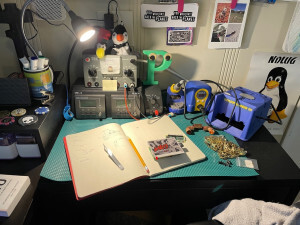 A desk with a soldering iron, digital oscilloscope, and a variety of miscellaneous items, lit up nicely by a small lamp