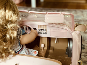 A doll is seen from behind sitting in a pink doll car. The car is a convertible and the doll’s head sticks above the windshield. Centered in the view is the car’s dashboard, including the buttons for the car’s radio