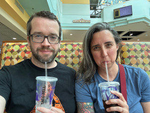 Jamie and Pete sitting in a food court, drinking from Taco Bell cups 
